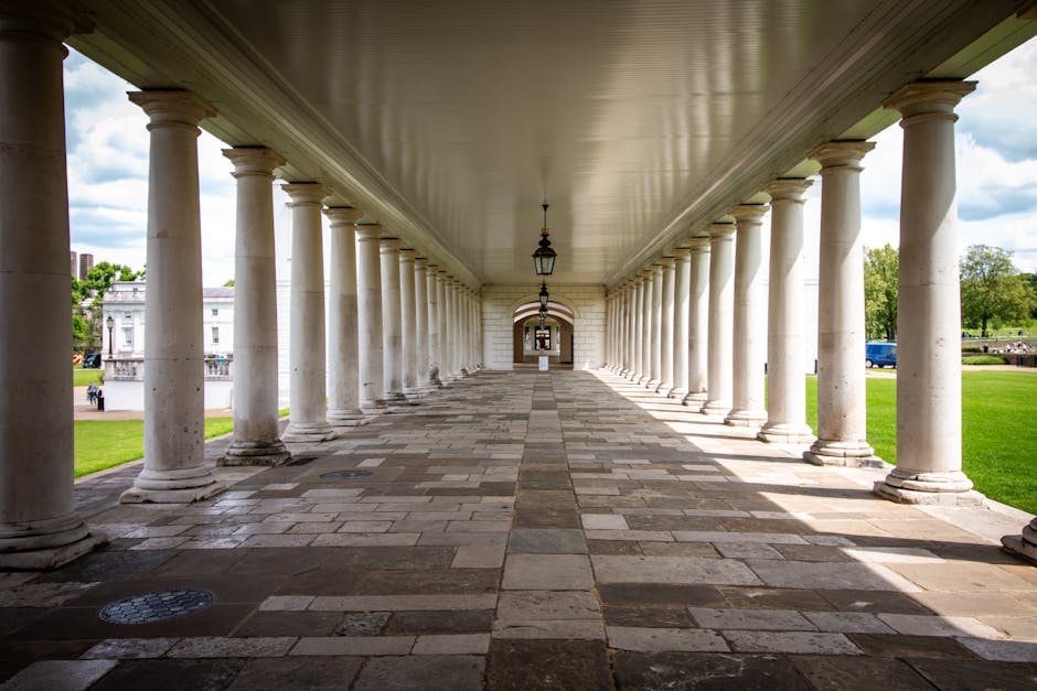 A long, covered outdoor walkway supported by a series of evenly spaced white stone columns, leading to an arched doorway at the far end. The ground is paved with rectangular stone tiles, some of which appear wet or shiny, and a black lantern-style light hangs from the ceiling between the columns. On either side of the walkway, there are well-maintained grassy areas with trees and a few parked vehicles visible in the background. The overhead ceiling is painted a neutral light color, and natural daylight illuminates the scene. This setting reflects a historic or classical architectural style, suitable for a public park or heritage site, with an emphasis on symmetry and visual depth, aligning with themes of home relocation and moving logistics as seen in house removals and furniture transport services offered by Man with Van Greenwich.