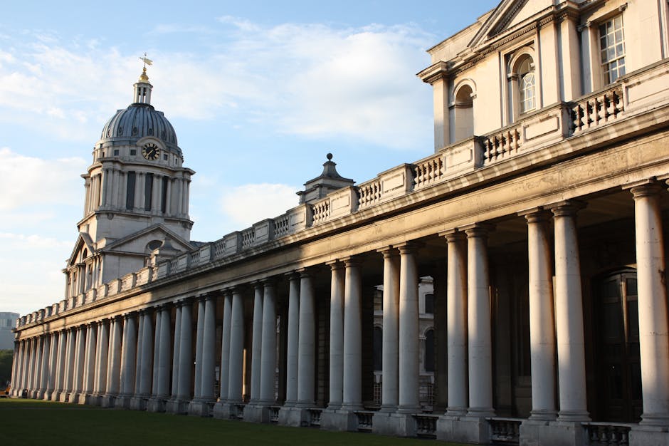 A long row of classical stone columns supporting an elegant, historic building with a large dome topped by a clock tower, situated outdoors on a clear day. The scene includes the building's exterior facade with detailed architectural features, and the sky above is partly cloudy with soft sunlight highlighting the stonework. In the foreground, inside a property, a man from Man with Van Greenwich is engaged in a home relocation process, carrying a cardboard box wrapped in plastic, indicating packing and moving activities. Nearby, there are several other cardboard boxes, some sealed with packing tape, placed on the floor and ready for transport. Additional packing materials such as bubble wrap and fabric blankets are visible, suggesting careful furniture transport. The Vandriving van is positioned outside, with its open back door ready for loading of furniture and boxes, supported by equipment like trolleys and straps seen in the area. This scene exemplifies professional packing, furniture transport, and loading processes involved in house removals in Greenwich, aligned with the services provided by Man with Van Greenwich.