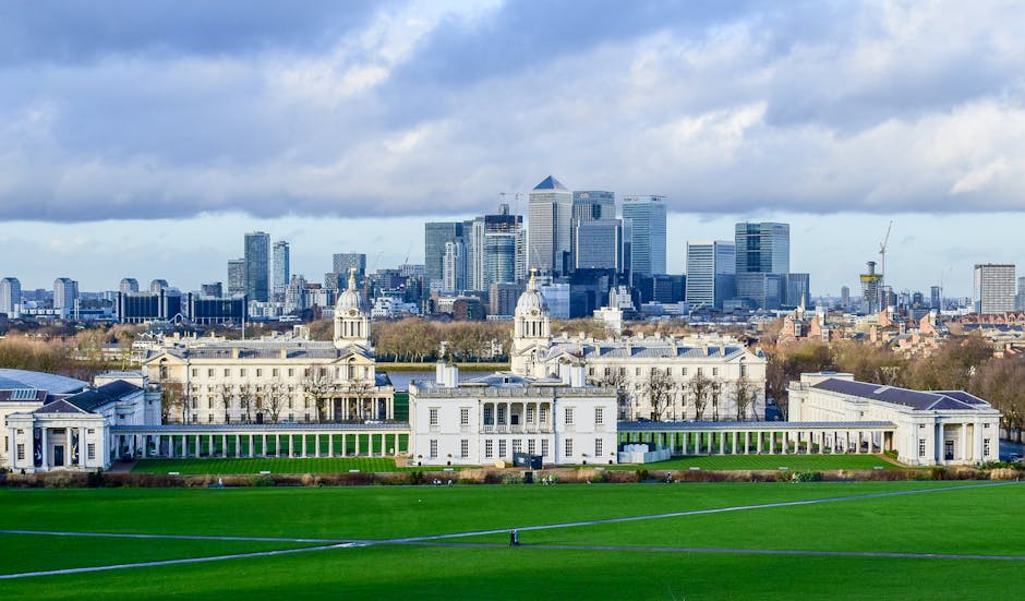 A wide-angle view of a large historic white building with two prominent towers and a long colonnade, situated in a lush green park with well-maintained grass and curved pathways in the foreground. Behind the building, a modern city skyline with tall glass skyscrapers under a partly cloudy sky is visible. The scene captures an urban landscape combining historic architecture and contemporary high-rises, indicative of a city like Greenwich, with clear evidence of a home relocation or moving process such as the presence of boxes or furniture in nearby areas. The overall setting suggests a professional removals service environment, with the backdrop emphasizing the area's blend of old and new structures.