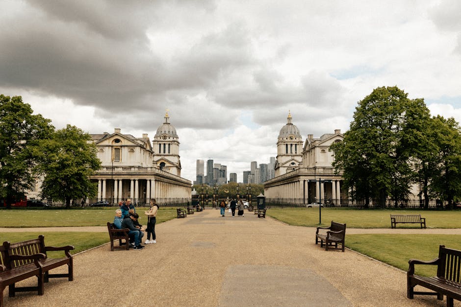 A wide view of a park with a gravel pathway leading towards two symmetrical, historic white stone buildings with domed roofs and classical columns, situated on either side of the path. Several trees are visible around the buildings, providing greenery against the cloudy sky. In the foreground, there are multiple wooden benches along the pathway, with some people sitting and conversing, while others stand nearby, possibly engaging in packing or planning a home relocation. A few individuals are walking further down the path towards the central part of the park, which features more benches, trash bins, and a distant cityscape with modern high-rise buildings. The scene suggests a transitional environment suitable for loading or unloading furniture and packing materials during house removals, with the presence of a van or vehicle not visible but implied, emphasizing the importance of efficient logistics by a professional removals service such as Man with Van Greenwich.