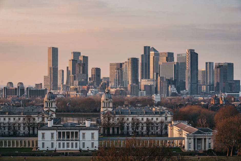 A wide-angle view of a large historic white building with two prominent towers and a long colonnade, situated in a lush green park with well-maintained grass and curved pathways in the foreground. Behind the building, a modern city skyline with tall glass skyscrapers under a partly cloudy sky is visible. The scene captures an urban landscape combining historic architecture and contemporary high-rises, indicative of a city like Greenwich, with clear evidence of a home relocation or moving process such as the presence of boxes or furniture in nearby areas. The overall setting suggests a professional removals service environment, with the backdrop emphasizing the area's blend of old and new structures.
