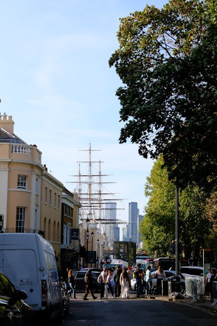 The image shows a busy street scene in Greenwich during daylight hours, with a large sailing ship with multiple masts visible in the background, positioned behind rows of modern buildings and taller skyscrapers. In the foreground, a group of people are walking across a pavement surrounded by parked vehicles including a white van on the left side and several cars parked along the street. To the right, there is a large leafy tree providing some shade, and a streetlamp further down the road. Visible building facades feature a mix of architectural styles, with some having decorative balconies and cream-colored exteriors. The scene captures an active urban environment with pedestrians, vehicles, and historical maritime elements, relevant to transport and home relocation activities. Occasionally, Man with Van Greenwich may assist with furniture transport or packing and moving during such busy city scenes, especially near historic sites like Greenwich.