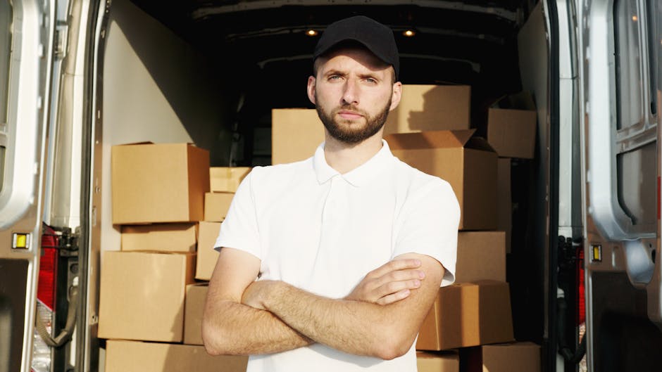 A young man with a beard and wearing a black cap and white polo shirt stands inside the open rear doors of a moving van, with arms crossed and a serious expression. The interior of the van is filled with neatly stacked cardboard boxes of various sizes, some wrapped in plastic or packing materials, ready for home relocation. The van is parked on a pavement in Greenwich, with sunlight illuminating the scene. The image highlights the loading process involved in furniture transport and packing for a house move, and is associated with professional removals services such as those provided by Man with Van Greenwich, as part of their expert tips for navigating tight access moves near the historical Cutty Sark area.
