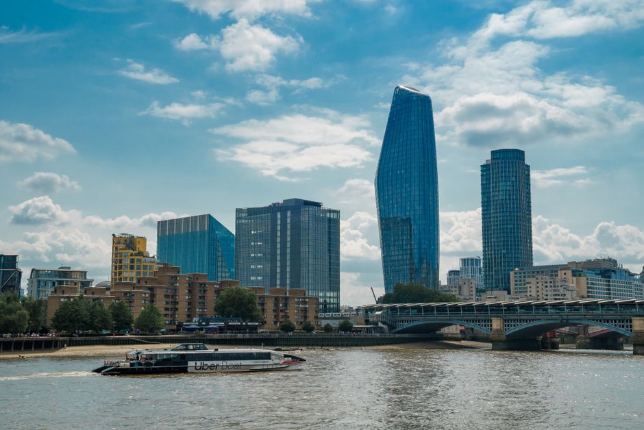 A city skyline featuring modern high-rise buildings with glass facades along the riverside, including a uniquely shaped curved skyscraper, with a bridge spanning the water and a black Uber boat traveling across the river. In the foreground, the river’s surface reflects the sky and nearby structures, with some greenery and low-rise buildings lining the waterfront. The scene appears during daytime with partly cloudy skies, providing natural lighting that highlights the reflective surfaces of the buildings. This urban landscape likely depicts an area undergoing professional house removals or furniture transport, with the possibility of loading or unloading occurring nearby, supported by the surrounding infrastructure. The image is consistent with a relocation service environment, emphasizing the logistical process of moving through cityscapes such as Greenwich or Deptford, as referenced in the services page for Man with Van Greenwich.