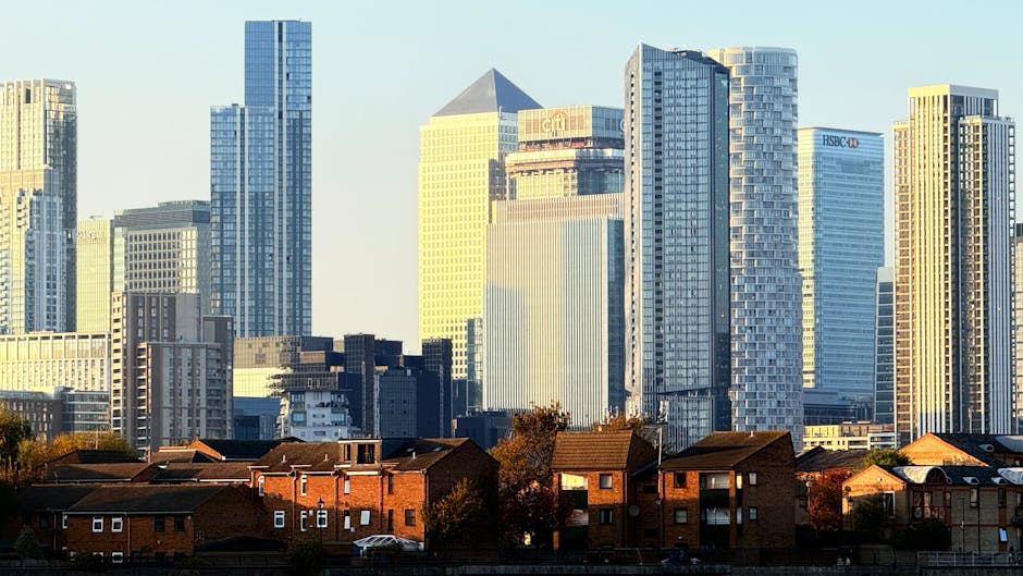 A daytime view of a city skyline featuring tall modern office skyscrapers with glass facades reflecting sunlight, situated behind a row of red-brick residential houses with small gardens and rooftops. The scene shows a contrast between the contemporary high-rise buildings in the background and the traditional houses in the foreground, set in Greenwich. The image captures the urban environment relevant to home and office removals, with the skyscrapers indicating a business district and the residential properties suggesting moving or relocation services nearby, as provided by Man with Van Greenwich.