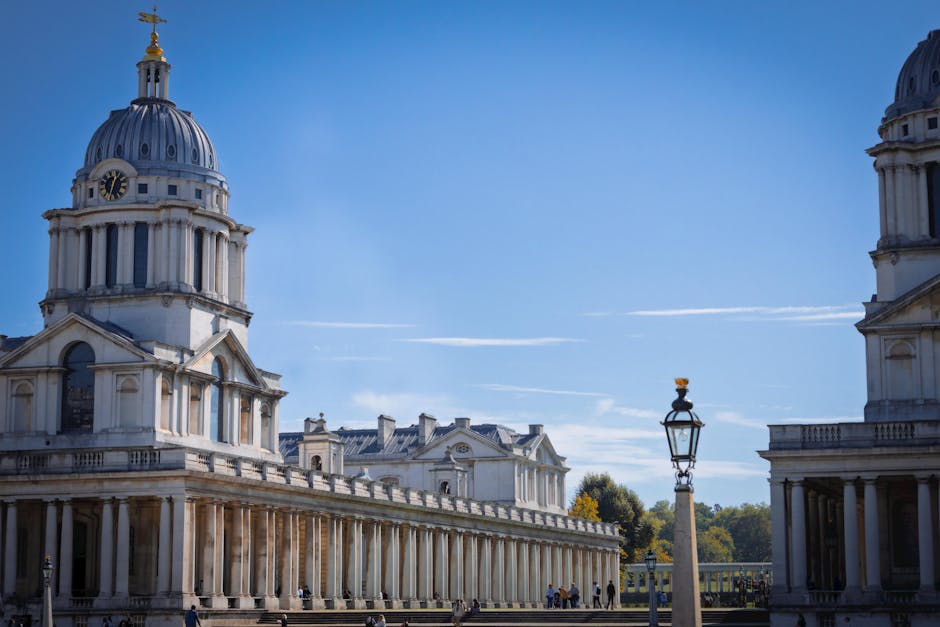 A clear daytime view of historic neoclassical buildings with domed roofs and ornate architectural details in Greenwich. The scene includes a lamppost with a decorative design, situated on a paved area with a few pedestrians visible in the background. The sky is blue with a few wispy clouds. This setting illustrates a recognizable landmark in Greenwich, which is a location often associated with house removals and moving services. The image captures the environment where furniture transport and packing for home relocation might take place, with the buildings serving as a backdrop to the logistical process of loading and unloading household items. Inside the property, packing materials like cardboard boxes and protective blankets may be used for safe transportation, and the loading process typically involves furniture trolleys or straps, with a removals van potentially visible nearby for efficient transport. Man with Van Greenwich offers home relocation services in this area, supporting customers with same-day removals and managing delays during the moving process.