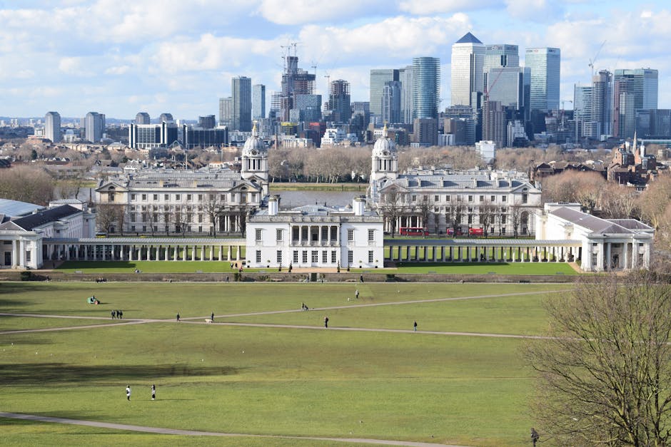 A wide-angle view of a historic white mansion with classical architecture, featuring two prominent central towers topped with small domes, set on a large, well-maintained green lawn with pathways and visitors walking around. Behind the mansion, a modern city skyline with tall glass skyscrapers and construction cranes is visible under a partly cloudy sky. In the foreground, the expansive park area includes scattered groups of people strolling, sitting, and enjoying the open space. The scene integrates elements of outdoor leisure, historic architecture, and urban development, reflecting a typical setting for home relocation and furniture transport logistics with local removals services like Man with Van Greenwich, capturing the transition between residential and city environments during a moving process.