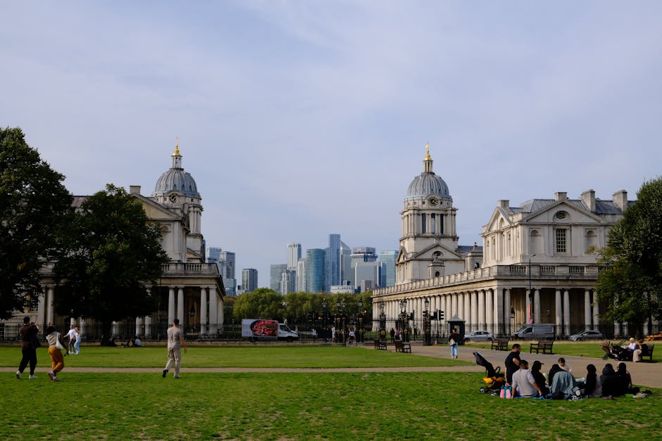 A wide view of Bolton's National Maritime Museum in Greenwich, London, showing the historic white stone buildings with classical architectural features, including domed roofs topped with golden finials, balconies, and columns, set in a green park area with several people relaxing, sitting on benches, and walking along the paved pathways. In the background, modern skyscrapers of the Canary Wharf financial district are visible under a partly cloudy sky. The scene captures a typical day of home relocation or furniture transport activities, with some individuals possibly assisting with packing or arranging items outside the museum, and others enjoying leisure activities. The outdoor setting features neatly maintained lawns, trees, and a busy urban atmosphere, reflecting the context of moving or packing services in Greenwich, as provided by Man with Van Greenwich.