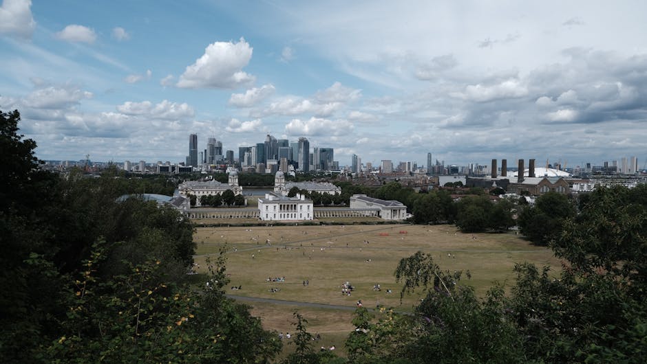 A wide-angle view of a historic white mansion with classical architecture, featuring two prominent central towers topped with small domes, set on a large, well-maintained green lawn with pathways and visitors walking around. Behind the mansion, a modern city skyline with tall glass skyscrapers and construction cranes is visible under a partly cloudy sky. In the foreground, the expansive park area includes scattered groups of people strolling, sitting, and enjoying the open space. The scene integrates elements of outdoor leisure, historic architecture, and urban development, reflecting a typical setting for home relocation and furniture transport logistics with local removals services like Man with Van Greenwich, capturing the transition between residential and city environments during a moving process.
